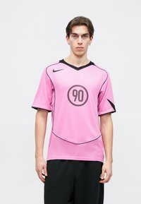 Ροζ φανέλα Nike με μαύρο V-λαιμό, κοντά μανίκια και στρογγυλό λογότυπο με το "90" στο κέντρο. Υφή υφάσματος και αντίθετες μαύρες λεπτομέρειες.