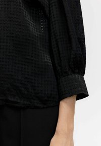 Top nero con motivo testurizzato di triangoli in rilievo, dotato di scollatura rotonda e polsini elasticizzati. Il materiale appare liscio e leggermente lucido.