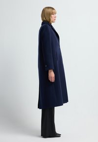 Marineblauer Wollmantel mit geradem Design, großem Reverskragen, doppelter Knopfleiste und seitlichen Schlitzen, kombiniert mit schwarz gestreiften Hosen.