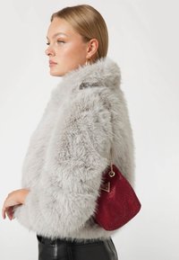 Giacca in pelliccia sintetica grigia con texture voluminosa, abbinata a una piccola borsa a rete rossa con dettagli metallici e una tracolla a catena.