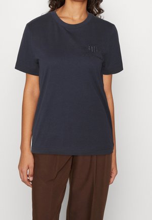 Marineblauw katoenen T-shirt met korte mouwen, ronde hals en reliëflogo op de linkerborst. Gecombineerd met losse, bruine broek.