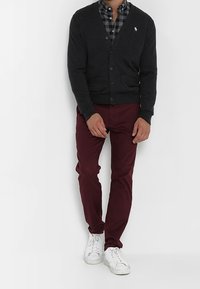 Donkergrijze cardigan met knopen over een zwart-wit geblokt shirt, gecombineerd met bordeauxkleurige slim-fit broek en witte sneakers.