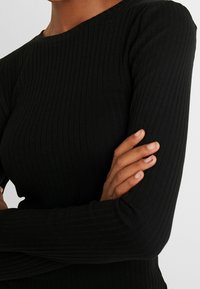 Svart ribbad långärmad topp med rund halsringning. Tyget ser mjukt ut med vertikal textur. Armarna korsade, vilket visar den figurnära designen.