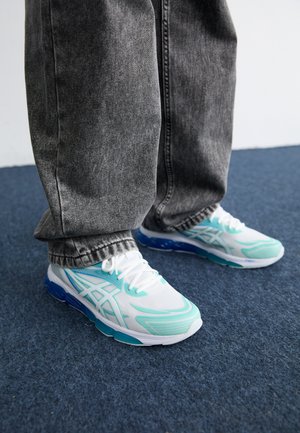 White athletic shoes with teal accents and blue soles, featuring a lightweight design and textured surface, worn with gray jeans.
