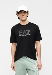 Zwart T-shirt met korte mouwen met wit "EA7 Emporio Armani" logo. Model draagt lichtgroene joggers, die een ontspannen pasvorm en zachte textuur tonen.