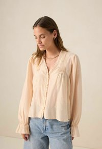 Blouse légère de couleur pêche avec un col volanté, des plis et des manches longues avec des poignets froncés. Texture de tissu lisse.