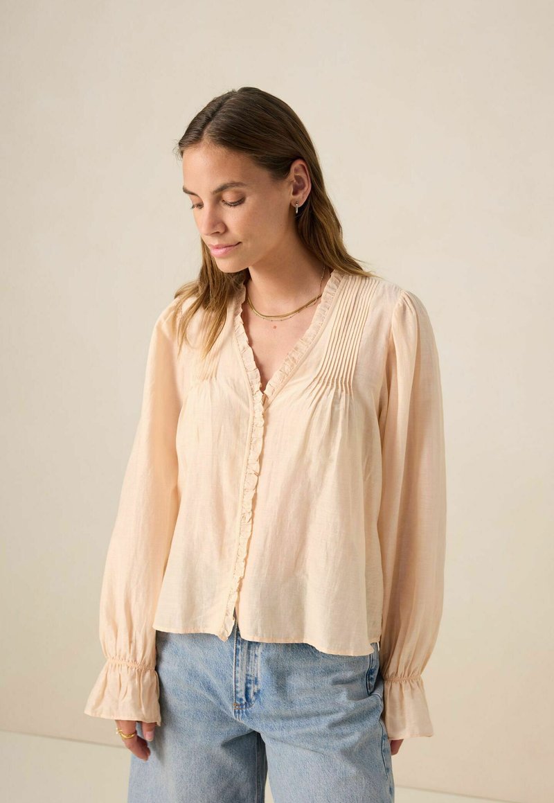 Blouse légère de couleur pêche avec un col volanté, des plis et des manches longues avec des poignets froncés. Texture de tissu lisse.