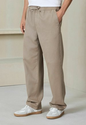 Man wearing loose beige drawstring pants with side pockets and white sneakers, standing indoors on a light floor.