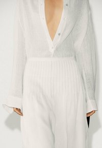 Hvid linnedskjorte med dyb V-udskæring, knappet front og lange ærmer. Den nederste del har diskrete lodrette striber. Let og tekstureret stof.