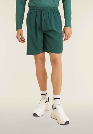 Shorts verts avec une ceinture élastique, fermeture à cordon, et une texture lisse. Associés à des baskets blanches et des chaussettes à motifs.