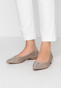 Ballerines en suède beige avec un capuchon en vernis brillant, présentant une forme pointe et un petit talon en bois, à côté d'un pantalon blanc longueur cheville.