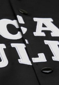 Camisa negra de tela con letras blancas bordadas y botones negros brillantes, con una textura suave y detalles de costura cuidados.