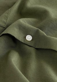 Tessuto verde oliva con una texture liscia, caratterizzato da un bottone bianco e cuciture a contrasto. Ingrandimento del dettaglio del tessuto.