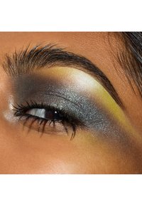 Primo piano di un occhio con ombretto grigio scuro e shimmer metallico, con un accento giallo e un sopracciglio ben curato sopra.