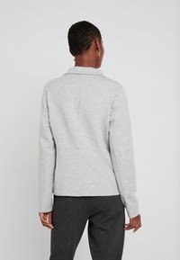 Šedé, texturované sako s límcem, dlouhými rukávy a zadními záševky; v kombinaci s tmavě šedými, na míru střiženými kalhotami.