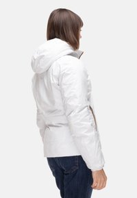 Giacca imbottita bianca con cappuccio, caratterizzata da una texture liscia e dettagli con cerniere arancioni. Indossata con jeans di denim blu, mostrata da una vista laterale.