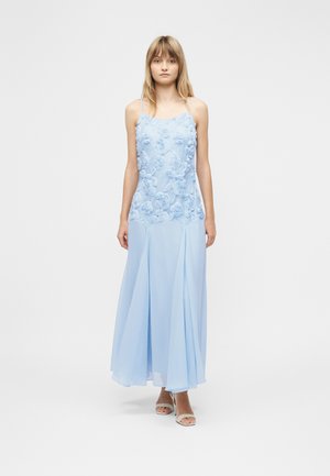 Frau trägt ein hellblaues, ärmelloses Kleid mit floralen Verzierungen am Oberteil und einem fließenden Rock, die vor einem schlichten weißen Hintergrund steht.