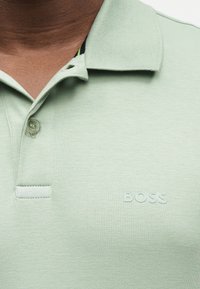 Gros plan d'un polo vert clair avec deux boutons, col et logo "BOSS" en relief sur la poitrine.