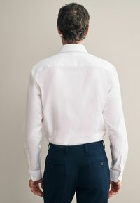 Weißes Langarmhemd mit klassischem Kragen, ausgestattet mit knöpfbaren Manschetten und einer glatten Textur, kombiniert mit dunkelblauen Hosen mit seitlichen Taschen.