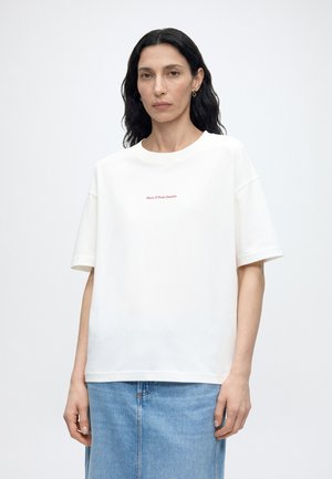 Γυναίκα με μακριά σκούρα μαλλιά που φοράει λευκό κοντομάνικο T-shirt με κόκκινο κείμενο "Marc O'Polo Denim" και μπλε τζιν φούστα, στέκεται μπροστά σε μονόχρωμο φόντο.