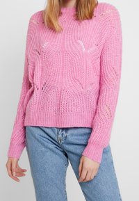Rosa Strickpullover mit einem strukturierten Wellenmuster und offenen Maschen, locker sitzend mit gerippten Bündchen, kombiniert mit hellen Jeans.
