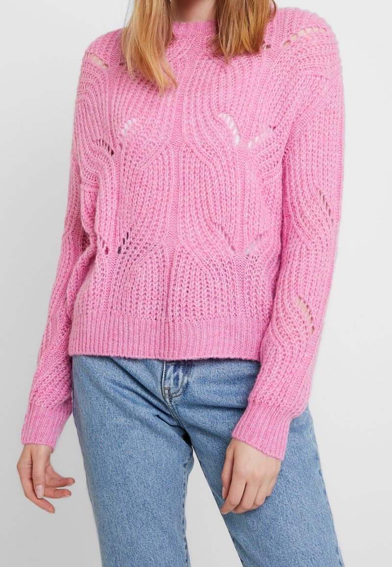 Rosa Strickpullover mit einem strukturierten Wellenmuster und offenen Maschen, locker sitzend mit gerippten Bündchen, kombiniert mit hellen Jeans.