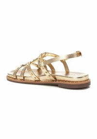 Sandalia plana de tiras metálicas doradas con tachuelas decorativas y correa en el tobillo. Textura suave y diseño elegante, que enfatiza la reflexión de la luz.