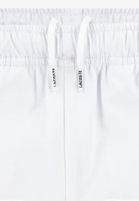 Bijele kupaće hlače s elastičnim pojasom i bijelim vezicama. Ukrašene crnim oznakama marke. Mekani, teksturirani materijal.