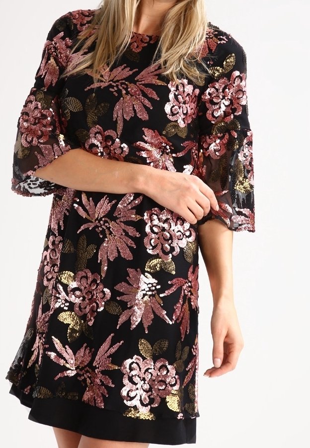 Robe noire avec des motifs floraux en sequins roses et dorés, avec des manches trois-quarts transparentes et un col rond, portée par une personne debout.