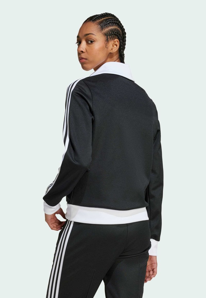 Chaqueta deportiva negra con acentos blancos y tres rayas a lo largo de las mangas. Cuello alto con puños y dobladillo acanalados. Textura de tela suave.