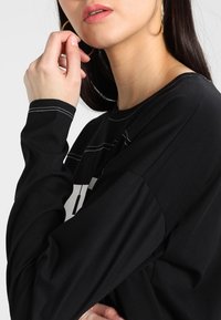 Svart långärmad tröja med kontrasterande vit sömnad, med en avslappnad passform och rundad halsringning. Modellen rör vid sitt ansikte med stora hoopörhängen.