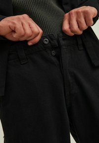 Jeans negros de algodón con cierre de botón, costuras visibles y una pretina texturizada. Manos agarrando la pretina en una vista en primer plano.