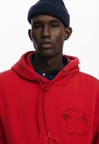 Červená mikina s kapucňou a textúrovanou grafiku medveďa. Obsahuje šnúrku na stiahnutie a pod ňou námornícky kockovaný golier.