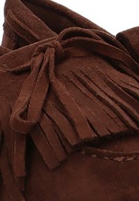 Mocasines de ante marrón con detalles de flecos y cierre de lazo. Textura suave con costuras visibles y diseño de punta redonda.