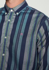 Polo s kratkimi rokavi z vertikalnimi črtami v mornarsko modri, teal in sivi barvi, z majhnimi zelenimi in rumenimi akcenti. Vključuje vezeno logotip na prsih.