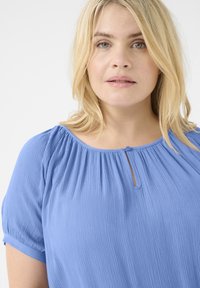 Blusa azzurra chiara con maniche corte e leggermente a sbuffo, tessuto testurizzato, scollatura arricciata e una piccola apertura a chiave con dettaglio di bottone.