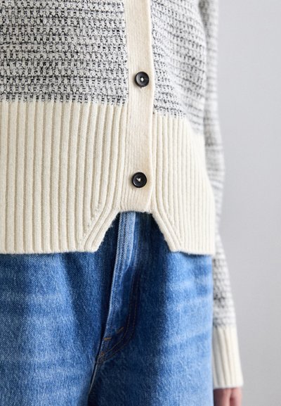 Strikket cardigan med en tekstureret grå øverste del og en ribbet cremefarvet nederste del. Har sorte knapper og en cropped skæring. Parret med blå jeans.