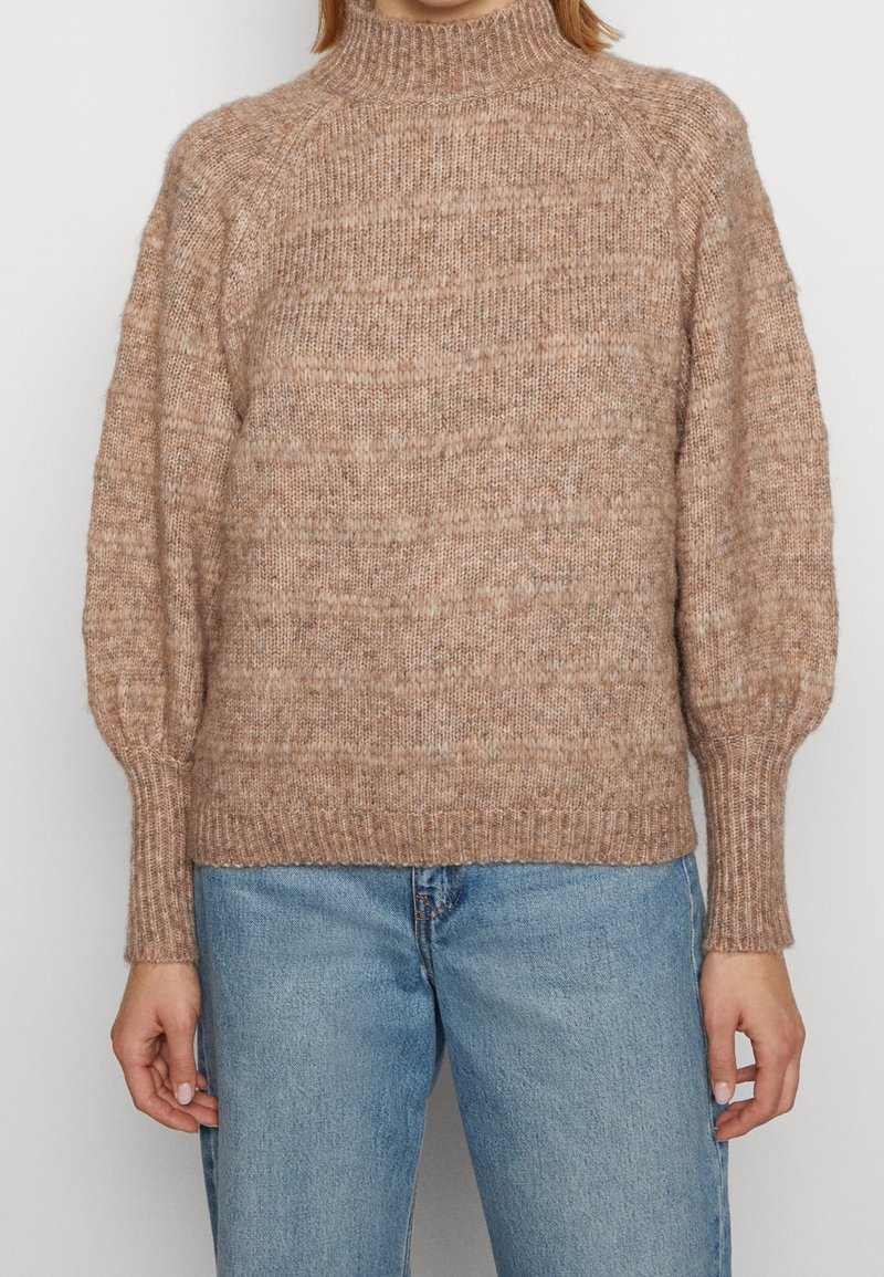 Pull en maille de couleur beige doux, col montant, poignets côtelés et coupe décontractée. Présente un tissu texturé avec des variations subtiles de couleur.