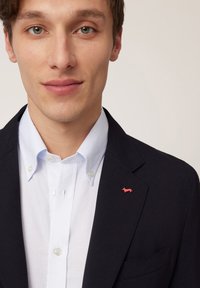 Mornarsko plava jakna s reverima preko bijele košulje s gumbima koja ima svijetloplavu ovratnik. Istaknuti crveni izvezeni logotip psa na jakni.