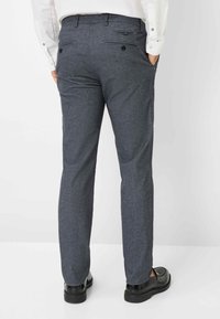 Grijze op maat gemaakte broeken van gestructureerde stof, met twee achterzakken met knopen, gedragen met zwarte schoenen.