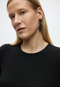 Ξανθιά γυναίκα με μαύρο τοπ που φοράει ένα μεγάλο συνθετικό σκουλαρίκι σε μαύρο και κόκκινο χρώμα, πάνω σε απλό φωτεινό φόντο.