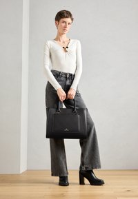 Black leather handbag with structured design, top handles, and zipper closure. Model wears a white long-sleeve top and gray wide-leg jeans.
