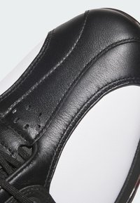 Czarny skórzany but sportowy z bliska, z gładką fakturą, perforowanymi detalami i białą podeszwą. Sznurowadła są czarne, widoczne szwy.