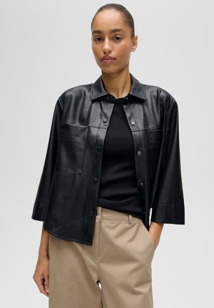 Frau mit zurückgebundenem Haar, die eine schwarze, knöpfbare Jacke aus Kunstleder, ein schwarzes Oberteil und beige Hosen trägt und eine Hand in der Tasche hat.