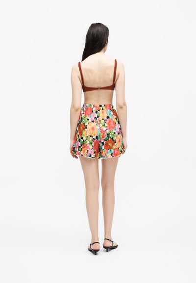 Γυναίκα που στέκεται με την πλάτη γυρισμένη, φοράει καφέ μπικίνι πάνω μέρος, σορτς ψηλόμεσα με λουλουδάτο μοτίβο και μαύρα σανδάλια με χαμηλό τακούνι.