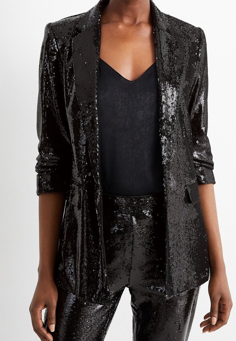 Blazer noir à sequins et pantalon assorti portés sur un top noir à col en V, avec les manches du blazer retroussées jusqu'aux coudes.
