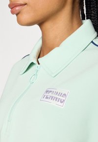 Piparu zaļš polo krekls ar rāvējslēdzēju, ar baltu logo tagu, uz kura rakstīts "TENNIS", un smalku zilu akcentu uz apkakles.