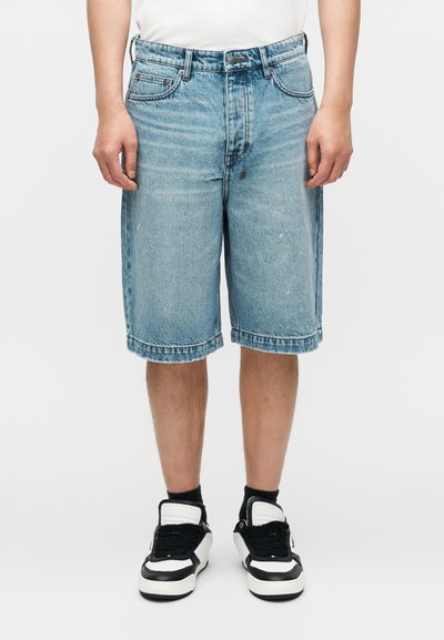 Shorts en denim bleu à coupe ample, longueur à mi-cuisse, design à cinq poches avec un léger effet délavé. Associés à des baskets noires et blanches.