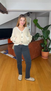 Νεαρή γυναίκα που στέκεται μέσα σε εσωτερικό χώρο φορώντας ένα κρεμ ζακέτα, μαύρο παντελόνι, μπεζ παπούτσια, με ένα μεγάλο φυτό και πορτοκαλί καναπέ στο φόντο.