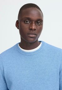 Pull léger bleu avec un tricot texturé, encolure ronde et une couche intérieure blanche au niveau du col. Gros plan montrant le détail du tissu et l'ajustement.